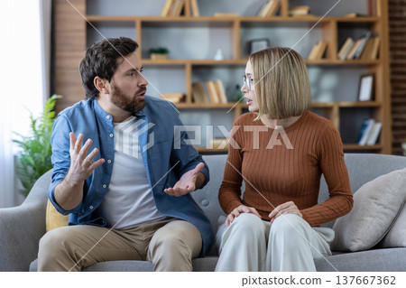Young couple is having a serious argument and gesturing while sitting apart on a sofa in their living room, indicating relationship problems and communication breakdown Young couple is having a serious argument and gesturing while sitting apart on a sofa in their living room, indicating relationship problems and communication breakdown 137667362