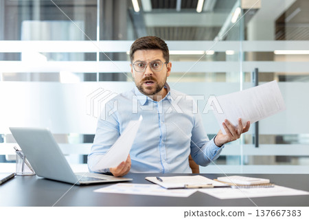Businessman wearing glasses looking surprised and frustrated holding documents at his office desk, indicating a problem, error, or unexpected news in his work 137667383