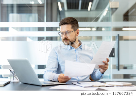 Professional man wearing glasses sitting at a desk in a contemporary office, carefully reviewing multiple business documents and paperwork next to a laptop, focusing intently on analysis and data 137667384