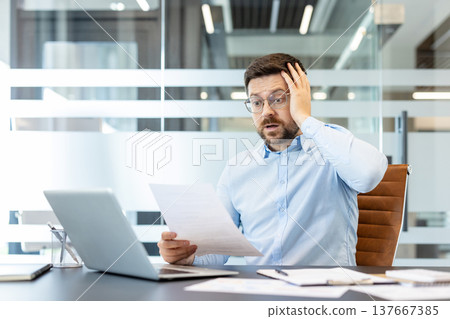 Businessman receiving stressful news while reviewing an official document, reacting with shock and worry about financial problems, debt, or an unforeseen crisis at work 137667385