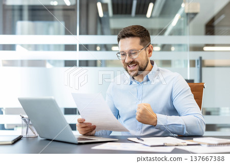 Businessman in modern office celebrates good news, clutching a document and clenching his fist with a joyful, triumphant smile at his desk, laptop nearby, dressed in professional attire 137667388