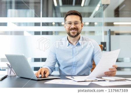 Smiling businessman sitting at desk in a modern office, working on his laptop while reviewing paper documents, representing concepts of productivity, success, and corporate professionalism Smiling businessman sitting at desk in a modern office, working on his laptop while reviewing paper documents, representing concepts of productivity, success, and corporate professionalism 137667389
