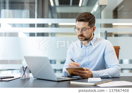Businessman in glasses and a light blue shirt actively taking notes in a spiral notebook while focusing on his laptop computer, working diligently at a modern office desk Businessman in glasses and a light blue shirt actively taking notes in a spiral notebook while focusing on his laptop computer, working diligently at a modern office desk 137667408