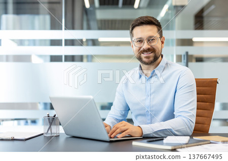 Young businessman sitting at desk, happily working on his laptop in a contemporary office setting, representing professional success, technology, and corporate environment Young businessman sitting at desk, happily working on his laptop in a contemporary office setting, representing professional success, technology, and corporate environment 137667495