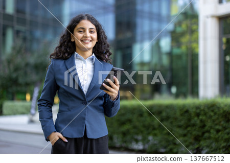 Young businesswoman with dark curly hair smiles confidently, holding a smartphone with one hand in pocket before a modern glass office building, symbolizing ambition and digital connectivity 137667512
