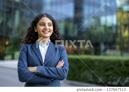 Confident indian businesswoman smiling with arms crossed, standing outdoors in front of a modern glass office building, symbolizing success, leadership, and professional achievement 137667513