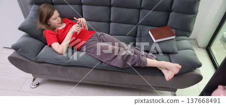 Child relaxes on a gray sofa while playing on a mobile device with a book nearby in a bright modern room 137668491