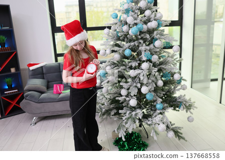 Festive girl in red shirt checks time while decorating a beautifully adorned Christmas tree in a cozy modern living room 137668558