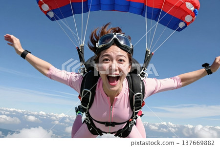 Thrilling skydiving portrait of a joyful young Asian woman in jumpsuit and goggles, soaring with arms outstretched under a colorful parachute amid blue skies and clouds. 137669882