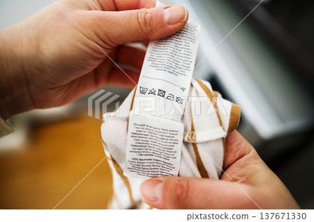Woman reading clothing label with care symbols and information on clothing near washing machine, closeup Woman reading clothing label with care symbols and information on clothing near washing machine, closeup 137671330