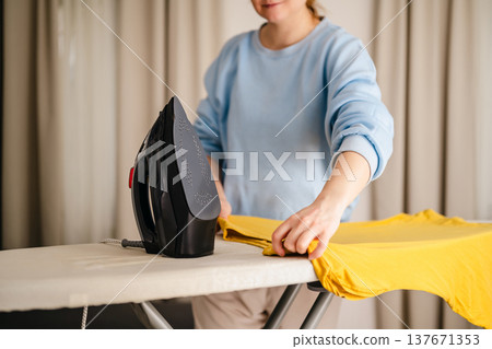 Woman in blue jamper, ironing an yellow t-shirts at home. Daily chores. Laundry day. Copy space Woman in blue jamper, ironing an yellow t-shirts at home. Daily chores. Laundry day. Copy space 137671353