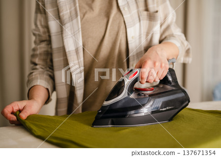 Home appliances. Woman ironing a t-shirts at home. Daily chores. Laundry day. Neutral background with copy space Home appliances. Woman ironing a t-shirts at home. Daily chores. Laundry day. Neutral background with copy space 137671354