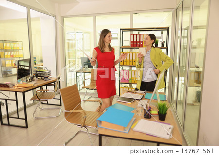 Bright office scene with two women engaged in lively discussion while surrounded by colorful files and modern workstations 137671561