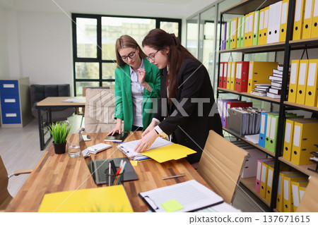 Two colleagues collaborate on a project in a bright modern office with organized shelves and a large window overlooking the city Two colleagues collaborate on a project in a bright modern office with organized shelves and a large window overlooking the city 137671615