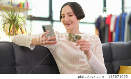 Cheerful woman enjoying online shopping while sitting comfortably on a cozy couch in a bright, modern living room Cheerful woman enjoying online shopping while sitting comfortably on a cozy couch in a bright, modern living room 137671665