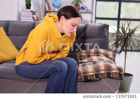 Woman in yellow hoodie sitting on couch, holding her stomach in discomfort during a quiet afternoon at home 137671681