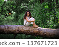 Little girl with curly hair sitting on a fallen tree trunk in a summer forest 137671723
