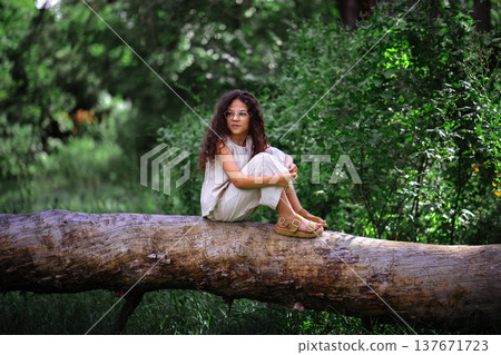 Little girl with curly hair sitting on a fallen tree trunk in a summer forest 137671723