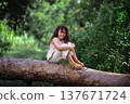 Happy little girl with curly hair smiling while sitting on a log in a sunny forest 137671724