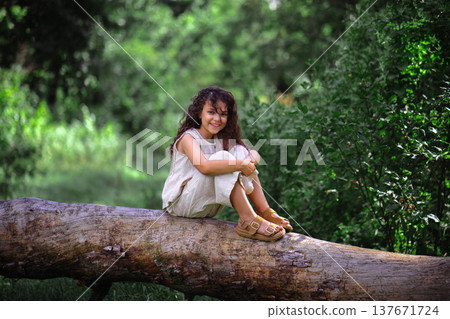 Happy little girl with curly hair smiling while sitting on a log in a sunny forest 137671724