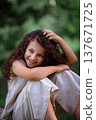 Adorable young girl with curly hair standing by a large tree in a green summer forest 137671725
