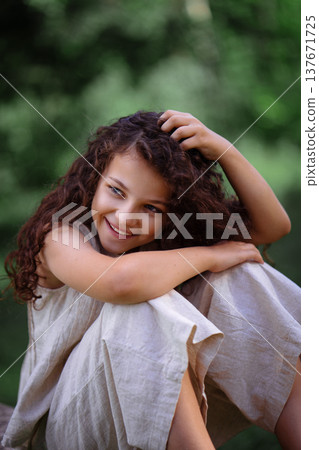 Adorable young girl with curly hair standing by a large tree in a green summer forest 137671725