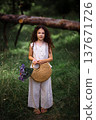 Little girl with long curly hair holding a woven straw bag with purple flowers in a forest path 137671726
