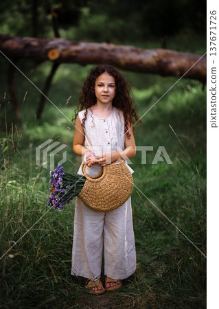 Little girl with long curly hair holding a woven straw bag with purple flowers in a forest path Little girl with long curly hair holding a woven straw bag with purple flowers in a forest path 137671726