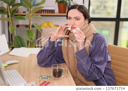 Sick woman sits at desk taking medicine and sipping coffee while wrapped in a cozy scarf during a cold morning at home Sick woman sits at desk taking medicine and sipping coffee while wrapped in a cozy scarf during a cold morning at home 137671754