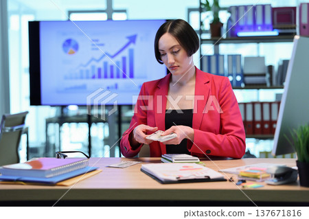 Focused professional reviewing financial reports and counting cash in a modern office space during the day Focused professional reviewing financial reports and counting cash in a modern office space during the day 137671816