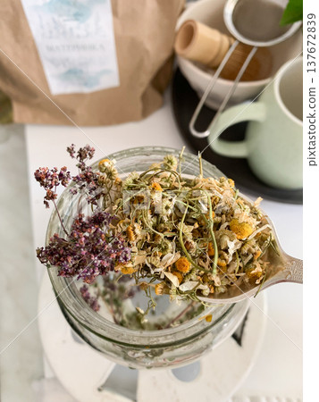 A person holds a spoonful of dried calendula and chamomile flowers, ready to make herbal tea. The kitchen has jars, a bag of herbs, and tea cups on a countertop A person holds a spoonful of dried calendula and chamomile flowers, ready to make herbal tea. The kitchen has jars, a bag of herbs, and tea cups on a countertop 137672839