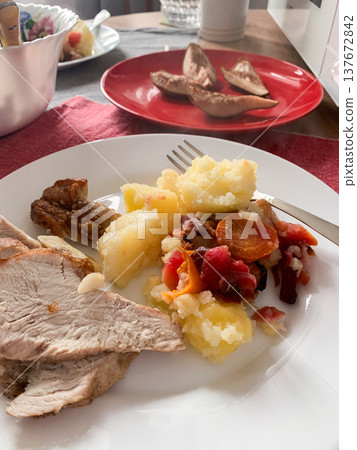 A plate holds sliced turkey meat, mashed potatoes, and a mix of colorful vegetables. In the background, baked potato wedges are served on a red plate. The setting is casual and inviting A plate holds sliced turkey meat, mashed potatoes, and a mix of colorful vegetables. In the background, baked potato wedges are served on a red plate. The setting is casual and inviting 137672842