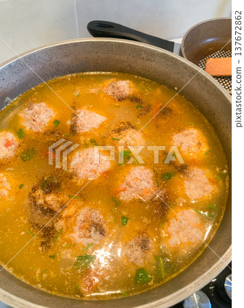 Meatballs are cooking in a pot of broth on the stove. The broth includes spices and vegetables. This shows meal preparation in a home kitchen 137672862