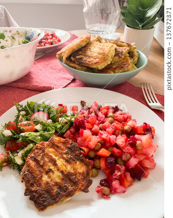 A meal features chicken cutlets, a fresh salad with chopped vegetables and quail egg. A colorful fruit drink is nearby on a table with a simple table setting in a kitchen A meal features chicken cutlets, a fresh salad with chopped vegetables and quail egg. A colorful fruit drink is nearby on a table with a simple table setting in a kitchen 137672871