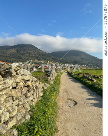 Oia, Galicia, Spain - March 27, 2025: Camino de Santiago rural path leading toward village houses, green hills with forested mountains. Camino de Santiago route landscape, rural village scenery Oia, Galicia, Spain - March 27, 2025: Camino de Santiago rural path leading toward village houses, green hills with forested mountains. Camino de Santiago route landscape, rural village scenery 137672879