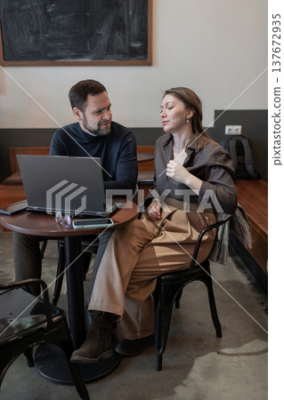 Business meeting at coffee shop, partners discussing project strategy across cups and plants. Business meeting, coffee shop, strategy, partners, teamwork. Business meeting at coffee shop, partners discussing project strategy across cups and plants. Business meeting, coffee shop, strategy, partners, teamwork. 137672935