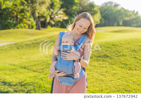 Mother using ergonomic baby carrier during a walk with her infant outdoors. Comfortable babywearing, active motherhood lifestyle, bonding, modern parenting, hands free care and everyday family routine 137672942