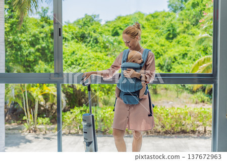 Woman vacuuming a room while carrying her baby in an ergonomic carrier. Modern motherhood, multitasking parent, home chores with infant, balance of childcare and housework concept. 137672963