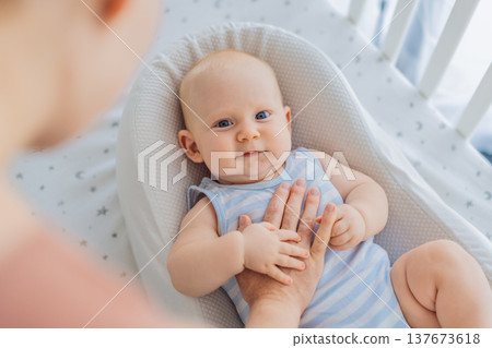 Mother gently sitting beside the crib and stroking her baby resting in a cozy cocoon nest. Tender intimate moment of care and trust at home. Motherhood, infant sleep and emotional bonding concept 137673618