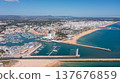 People visit Vilamoura marina in Portugal's Algarve region, enjoying boats and beach while the city skyline stands tall in the distance under clear blue sky Aerial 137676859