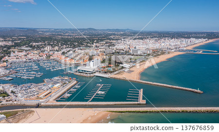 People visit Vilamoura marina in Portugal's Algarve region, enjoying boats and beach while the city skyline stands tall in the distance under clear blue sky Aerial 137676859
