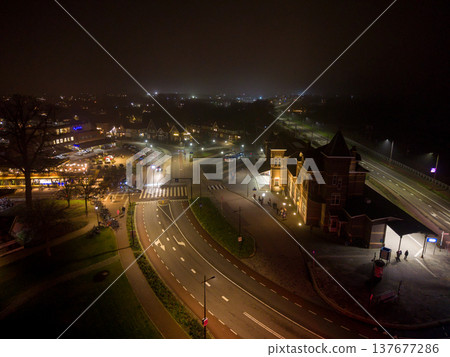 Aerial night view of the Kampen train station and surrounding city streets in the Netherlands. The scene features illuminated historic architecture, a bus terminal, and glowing street lights. Aerial night view of the Kampen train station and surrounding city streets in the Netherlands. The scene features illuminated historic architecture, a bus terminal, and glowing street lights. 137677286