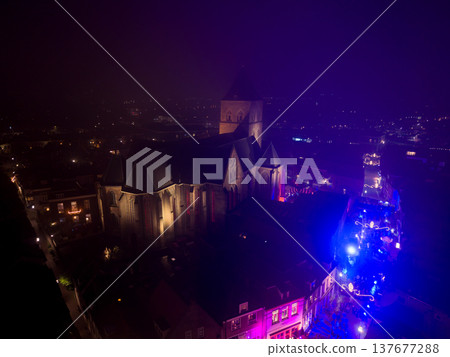 Aerial night view of the Nieuwe Toren tower in Kampen, Netherlands, illuminated in blue. The historic city is covered in mist, with festive Christmas market lights glowing in the streets below. Aerial night view of the Nieuwe Toren tower in Kampen, Netherlands, illuminated in blue. The historic city is covered in mist, with festive Christmas market lights glowing in the streets below. 137677288