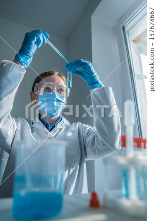 Young researcher in lab coat and safety goggles works with test tubes, focusing on precise measurement. Laboratory environment highlights innovation, science, and medical discovery 137678667