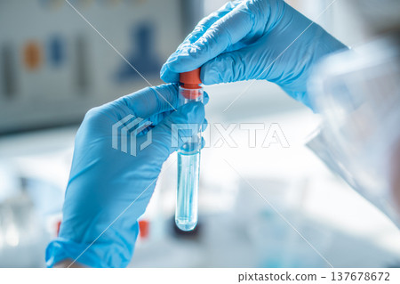 Closeup of gloved hands handling a test tube with clear solution in a bright scientific lab environment. Laboratory testing, diagnostics, and biotechnology research for medical advancements 137678672