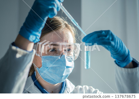 Female scientist wearing safety equipment carefully transfers liquid with pipette in laboratory setting. Professional researcher performs chemical analysis in bright, sterile environment 137678675