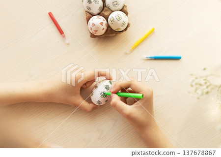 Children's hands use green marker to color simple flower designs on white eggs on table, with other eggs in tray and markers nearby, Easter, handmade Children's hands use green marker to color simple flower designs on white eggs on table, with other eggs in tray and markers nearby, Easter, handmade 137678867