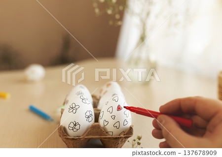 Close-up of person decorating Easter eggs with red marker. White eggs with hand-drawn hearts and flowers placed in an egg carton on table. Creative Easter preparation, DIY holiday decoration 137678909