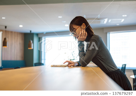 A woman wearing glasses is looking at a tablet in a cafe. A woman wearing glasses is looking at a tablet in a cafe. 137679297