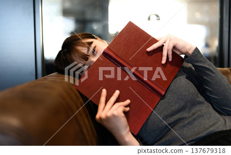 A woman sitting on a sofa with a book in front of her face. A woman sitting on a sofa with a book in front of her face. 137679318
